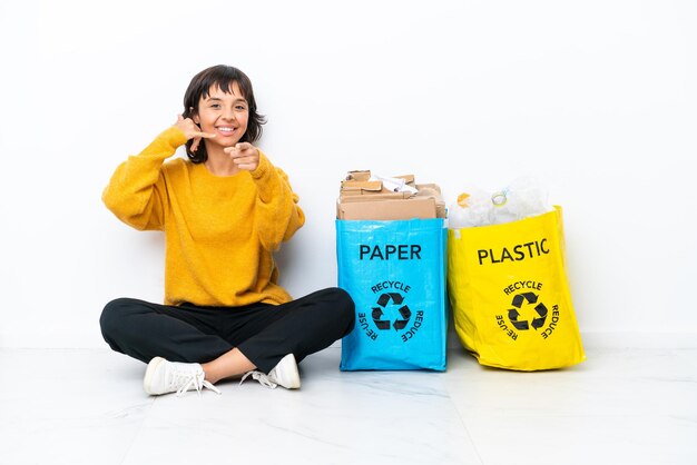 Jeune fille tenant un sac plein de plastique et de papier assis sur le sol isolé sur fond blanc faisant un geste de téléphone et pointant vers l'avant