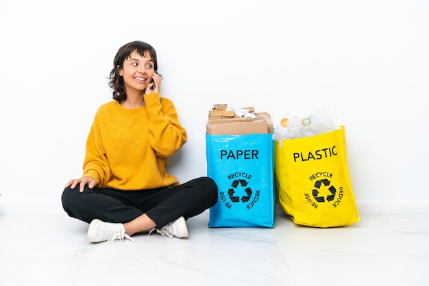 Jeune fille tenant un sac plein de plastique et de papier assis sur le sol isolé sur fond blanc en gardant une conversation avec le téléphone portable avec quelqu'un