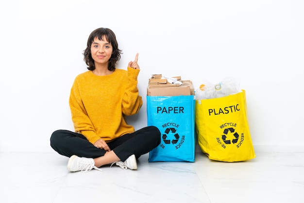 Jeune fille tenant un sac plein de plastique et de papier assis sur le sol isolé sur fond blanc pointant avec l'index une excellente idée