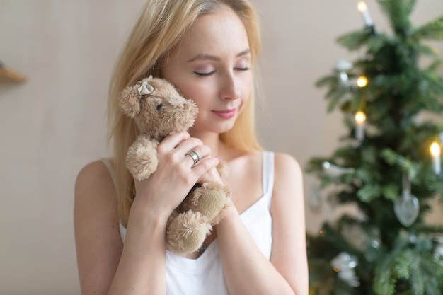 Jeune fille tendre en nuisette embrasse ourson avec arbre de Noël sur fond.