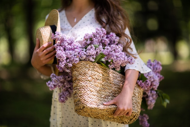 La jeune fille tient dans ses mains un panier en osier avec des fleurs. Panier avec des lilas. Fille et fleurs. Marchez avec un panier de lilas dans les mains. Fleuriste. Ambiance chaleureuse avec des fleurs fraîches et parfumées.