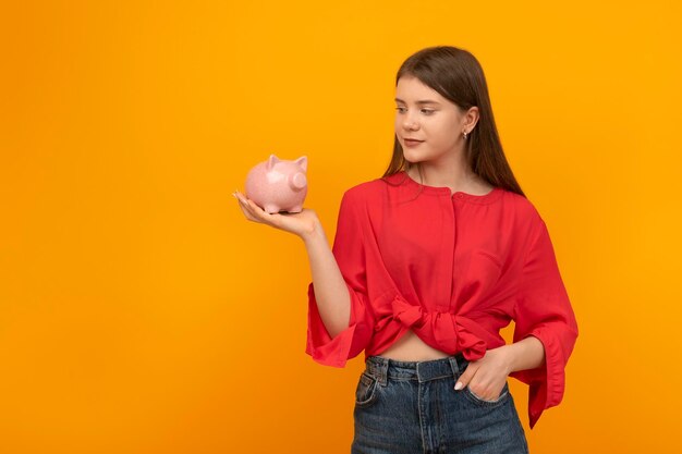 Une jeune fille tient une tirelire sur la paume sur fond orange vif Littératie financière Économiser de l'argent