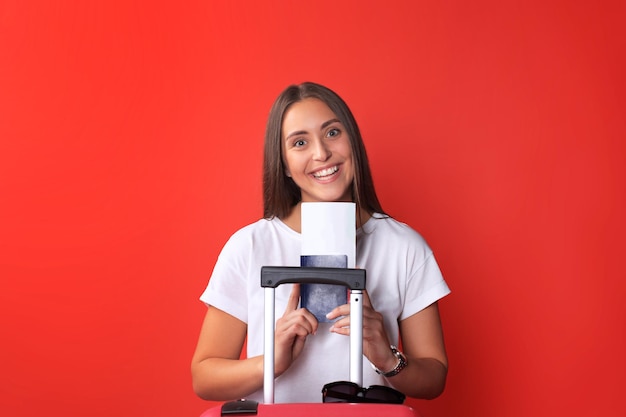 Jeune fille touristique en vêtements décontractés d'été, avec lunettes de soleil, valise rouge, passeport isolé sur fond rouge.