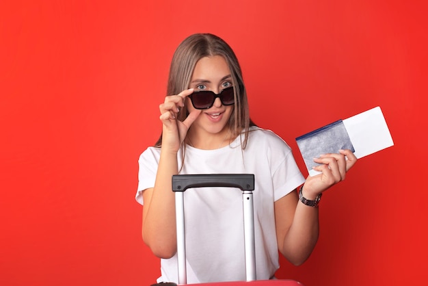Jeune fille touristique en vêtements décontractés d'été, avec lunettes de soleil, valise rouge, passeport isolé sur fond rouge.