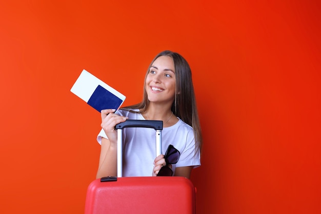 Jeune fille touristique en vêtements décontractés d'été, avec lunettes de soleil, valise rouge, passeport isolé sur fond rouge.