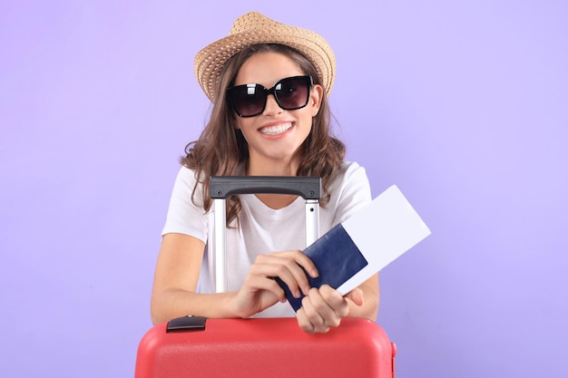 Jeune fille touristique en vêtements décontractés d'été, avec lunettes de soleil, valise rouge, passeport isolé sur fond violet