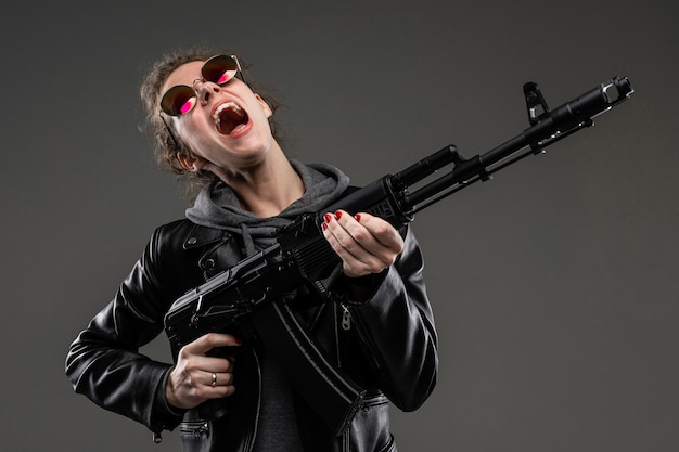 Jeune fille avec des traits du visage rugueux, des cheveux bruns, une manucure lumineuse, en vélo gris, une veste noire, avec des émotions violentes est titulaire d'un fusil d'assaut