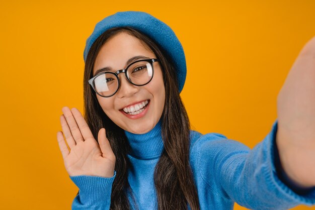 Jeune fille utilisant un téléphone portable montrant un geste de salut tout en faisant un appel vidéo sur fond jaune