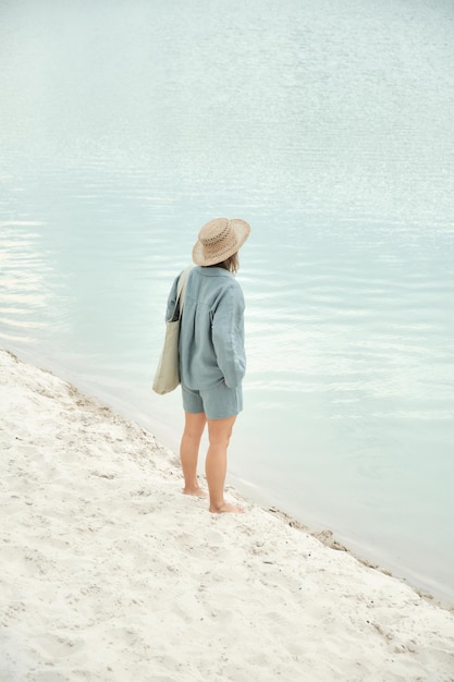 Jeune fille en vêtements de lin naturel et un chapeau de paille pieds nus sur une plage de sable blanc tenant un linge