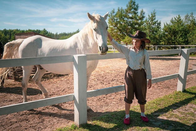 Jeune fille en vêtements vintage debout avec son cheval blanc brillant dans le paddock à la journée ensoleillée t...