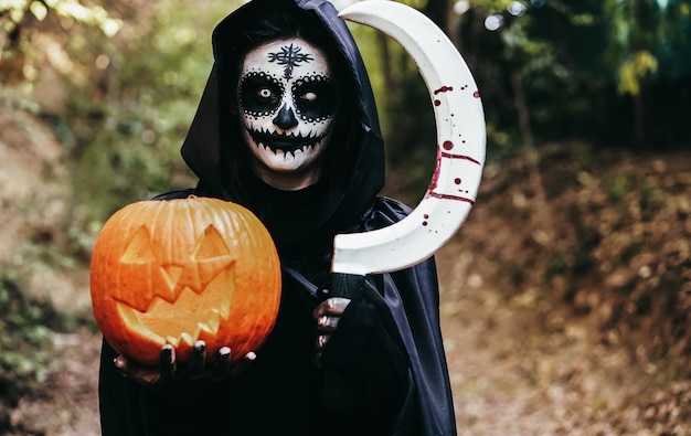 Jeune fille vêtue d'une mystérieuse robe d'Halloween tenant une citrouille effrayante et une faucille avec du sang dans les bois - Focus on face