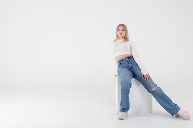 Une jeune fille vêtue d'un pull blanc et d'un jean est assise et pose sur un fond blanc isolé en studio. Concept de mode de vie des gens. Espace de copie pour copie