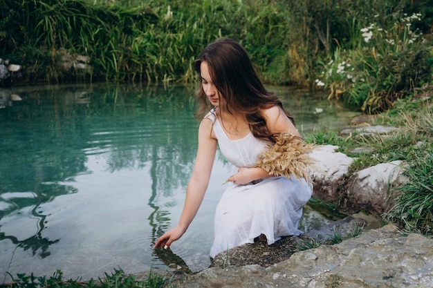 Jeune fille vêtue d'une robe blanche par une journée ensoleillée près du lac dans les bois. Fille dans un chapeau de paille sur les rives d'un lac forestier. Beau lac bleu au milieu de la forêt.