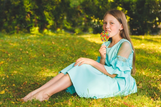 Une jeune fille vêtue d'une robe bleue se trouve sur l'herbe dans le parc avec une douce sucette