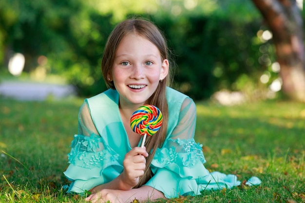 Une jeune fille vêtue d'une robe bleue se trouve sur l'herbe dans le parc avec une douce sucette