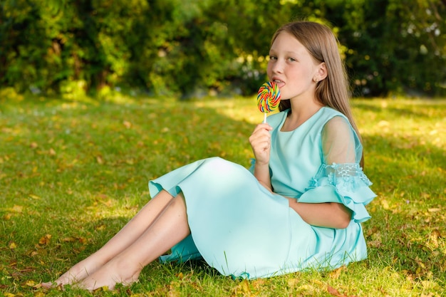 Une jeune fille vêtue d'une robe bleue se trouve sur l'herbe dans le parc avec une douce sucette