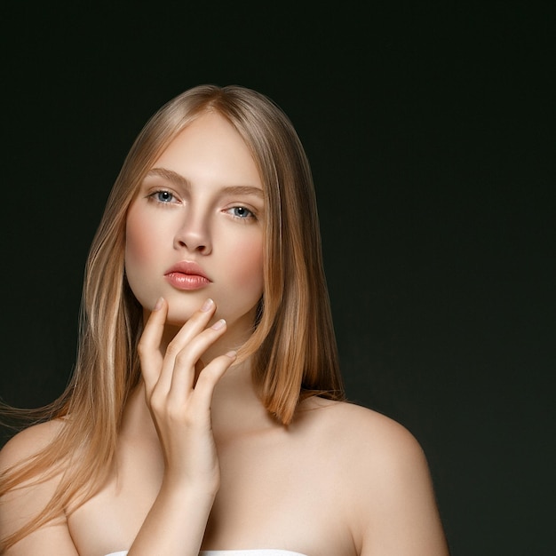 Jeune fille visage beauté peau portrait avec de longs cheveux blonds avec la main sur fond sombre. Prise de vue en studio.