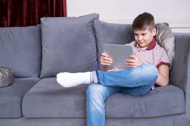 Jeune garçon adolescent beau à l'aide de tablette assis sur un canapé gris à la maison