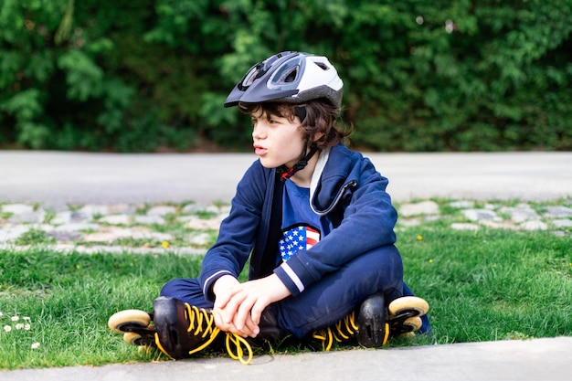 Jeune garçon adolescent sur patins à roulettes en casque assis sur l'herbe dans le parc, se reposer et se détendre. Notion de passe-temps.