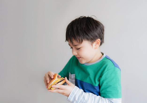 Jeune garçon en bonne santé mangeant un hamburger Un enfant tenant un cheeseburger et regardant avec un visage souriant Isolé Happy Kid ayant un hamburger de restauration rapide pour la collation La nourriture préférée des enfants