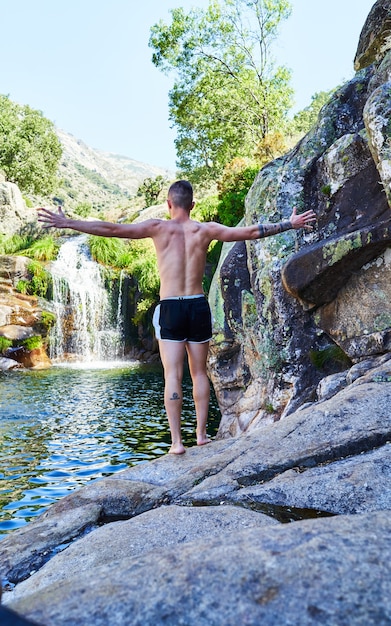 Jeune garçon avec les bras croisés derrière le dos sur un rocher en regardant la rivière avec cascade.