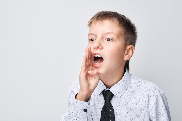 Jeune garçon en chemise et cravate criant ou criant avec la main couverte à la bouche à l'arrière-plan clair