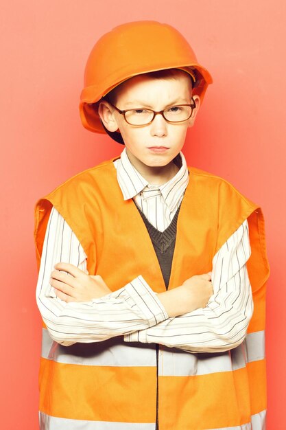 Jeune garçon constructeur mignon en uniforme orange et casque ou casque avec des lunettes sur un visage sérieux sur fond de studio rouge