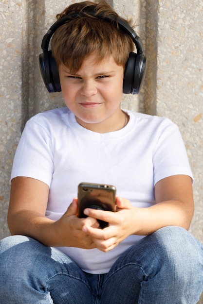 Le jeune garçon dans des écouteurs avec le smartphone écoute la musique en parc d'été