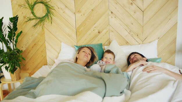 Jeune garçon drôle se réveille pendant que ses parents dorment le matin sur le lit dans leur maison