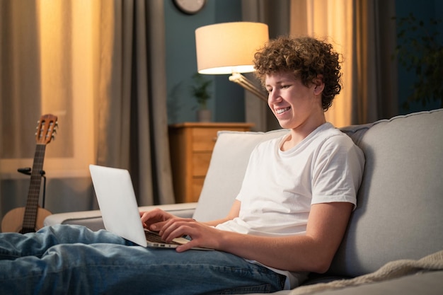 Un jeune garçon est assis dans le salon le soir et utilise un ordinateur portable