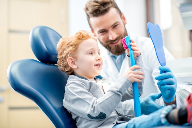 Jeune garçon excité assis sur la chaise tenant une grosse brosse à dents avec un médecin au cabinet dentaire