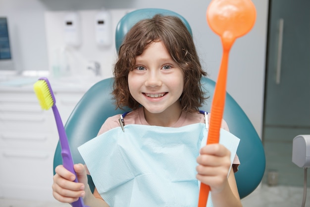 Jeune garçon excité souriant à la caméra, tenant un grand miroir dentaire et une brosse à dents au bureau du dentiste