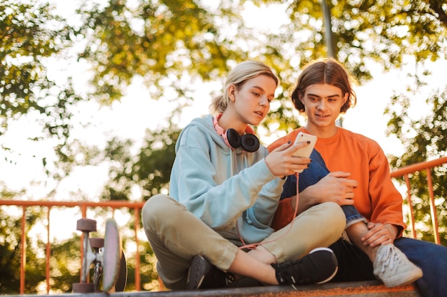 Jeune garçon et fille de patineur utilisant judicieusement leur téléphone portable tout en passant du temps ensemble au skatepark