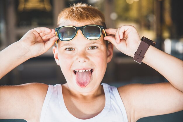 Jeune garçon à lunettes de soleil tire la langue.