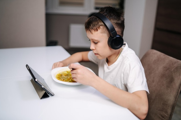 Jeune garçon mangeant et jouant sur une tablette avec un casque à la maison