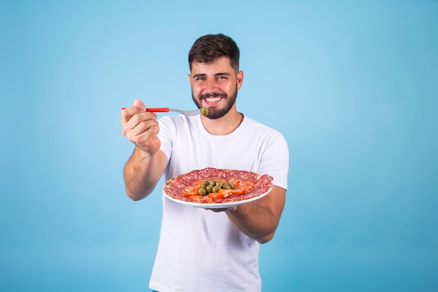 Un jeune garçon mangeant une portion de tomates et d'olives au salami tient une olive avec une fourchette et la montre à la caméra