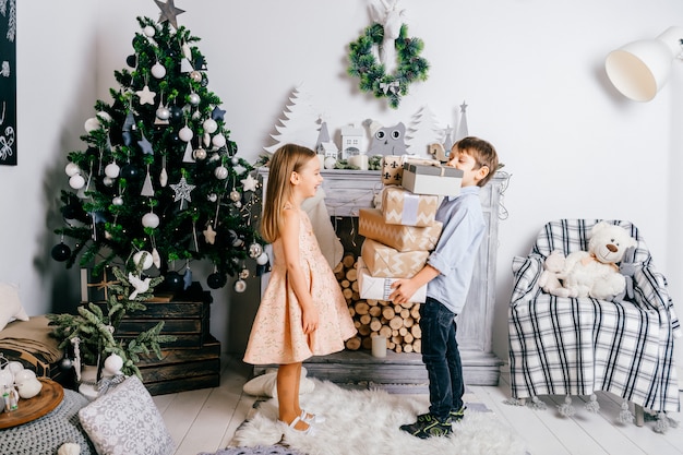 Jeune garçon présentant des coffrets cadeaux à une jolie petite fille dans la chambre avec arbre cristmas et cheminée. Vacances d'hiver.