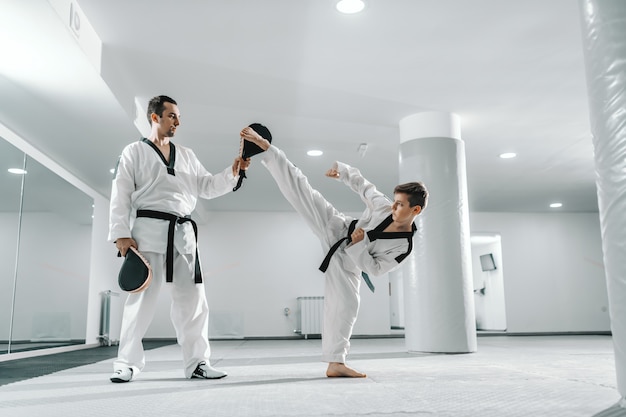 Jeune garçon de race blanche en dobok coups de pied pieds nus pendant que l'entraîneur tient la cible de coup de pied. Concept de formation de taekwondo.