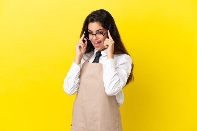 Jeune garçon de restaurant caucasien isolé sur fond jaune avec des lunettes et surpris