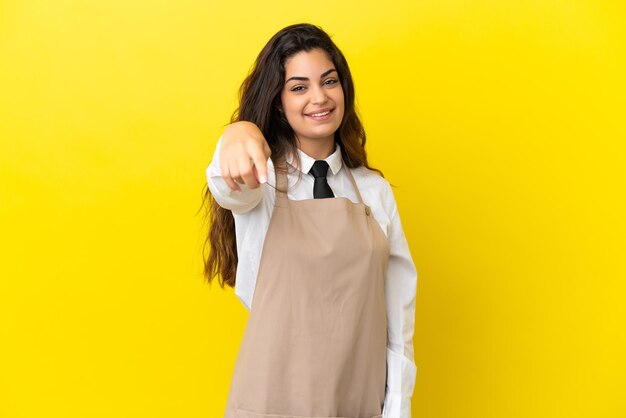 Jeune garçon de restaurant caucasien isolé sur fond jaune pointe le doigt vers vous avec une expression confiante