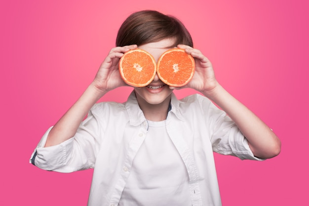 Jeune garçon se couvre les yeux avec une orange en tranches souriant à la caméra sur un mur rose