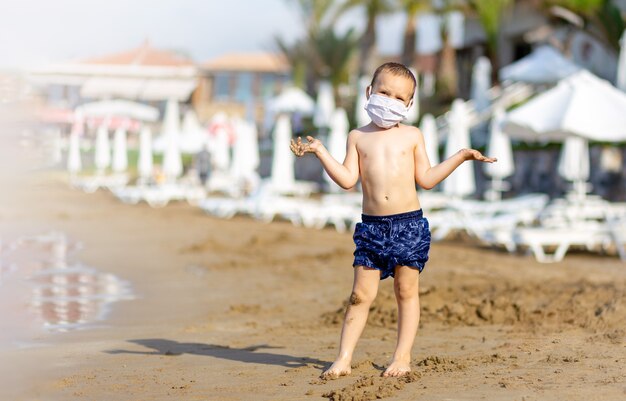 Jeune garçon en short de bain avec masque médical sur la plage se protégeant du virus corona, covid-19