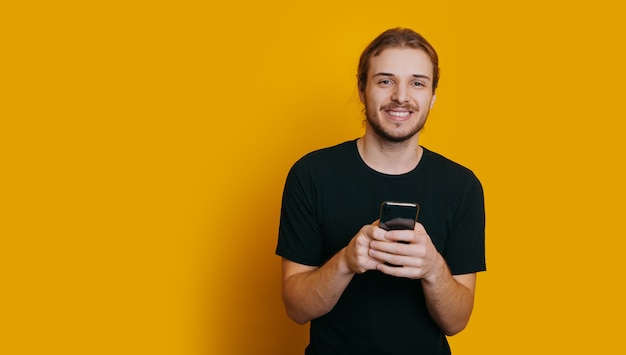 Un jeune garçon souriant avec barbe et cheveux longs tenant son téléphone