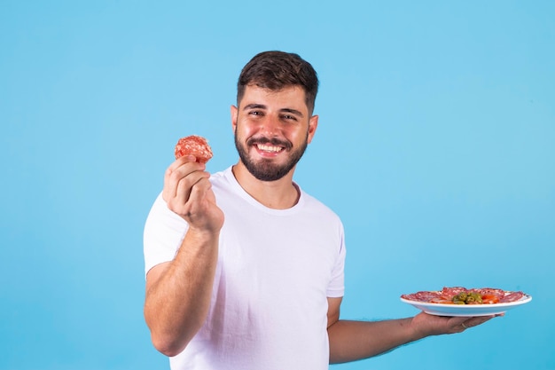 Jeune garçon tenant une assiette de salami et le mangeant sur fond bleu l'homme mange du salami
