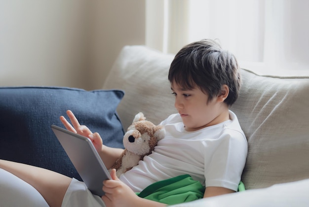 Jeune garçon utilisant une tablette jouant à un jeu sur Internet Enfant assis sur le canapé regardant ou parlant avec un ami en ligneEnfant se détendant dans le salon le matinEnfants avec le concept de la nouvelle technologie