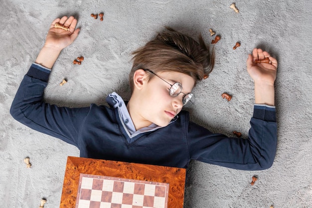 Jeune garçon vainqueur d'échecs dans des verres allongé sur le tapis autour de pièces d'échecs et échec et mat, vue de dessus