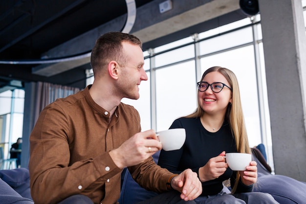 Une jeune gestionnaire réussie avec son patron est assise avec un ordinateur portable sur un fauteuil rembourré près de la fenêtre panoramique et boit du café. Femme et homme d'affaires travaillant sur un nouveau projet