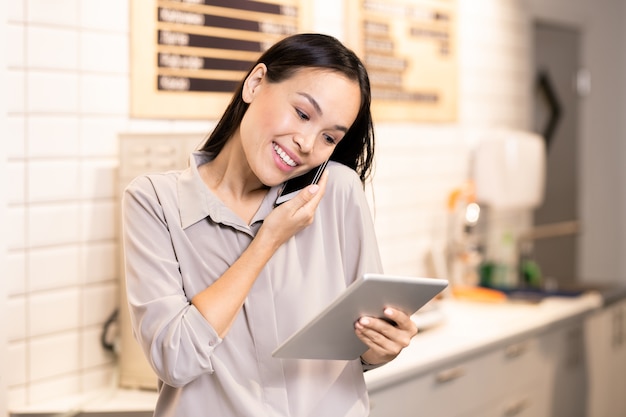 Jeune gestionnaire souriant d'un restaurant luxueux faisant défiler le menu en ligne sur tablette tout en consultant l'un des clients au téléphone