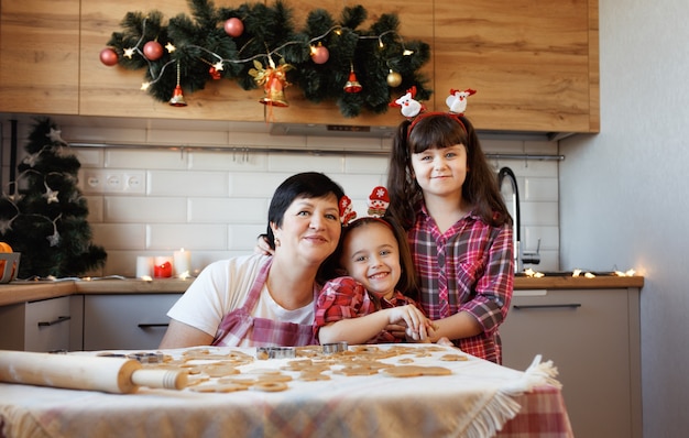 Une jeune grand-mère avec deux petites-filles se serre et rit dans la cuisine décorée pour les vacances de Noël.