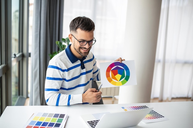 Jeune graphiste indépendant Guy travaillant avec un ordinateur portable et des échantillons de couleurs assis au bureau dans le bureau à domicile en choisissant la couleur gamma pour le nouveau projet de conception vue latérale avec espace de copie